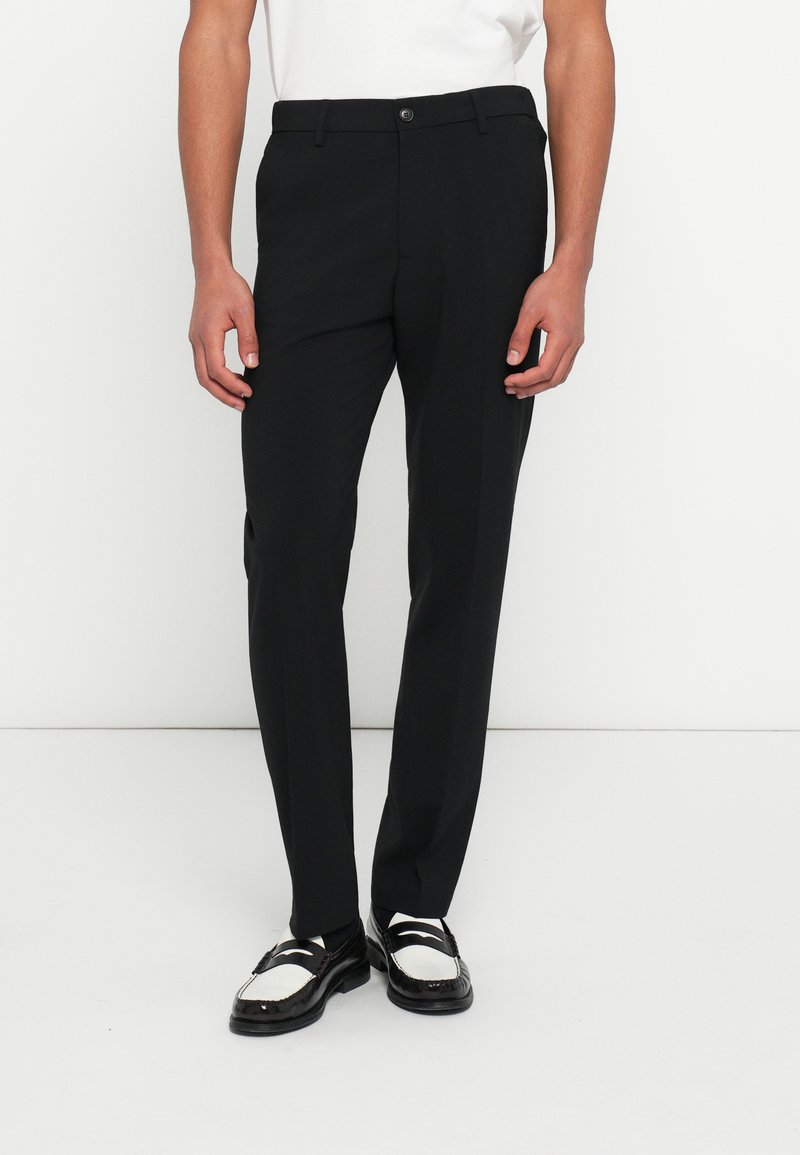 Zwarte op maat gemaakte broek met een rechte pijp, voorzien van een tailleband met knopen en zakken. Gecombineerd met zwart-witte loafers.