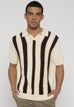 Mann trägt ein cremefarbenes Strickshirt mit kurzen Ärmeln, vertikalen dunkelbraunen Streifen und Kragen, steht mit den Händen in den Taschen einer khakifarbenen Hose.