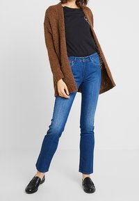 Cardigan marrón de punto sobre una camiseta negra, combinado con jeans rectos azules y mocasines negros. El cardigan presenta un diseño de frente abierto.