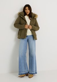 Chaqueta acolchada color verde oliva con capucha con borde de piel, presenta cierre delantero con botones a presión, bolsillos laterales y un diseño texturizado, combinada con vaqueros claros acampanados.