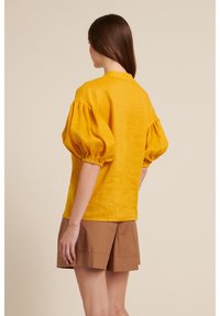 Blusa gialla in lino con maniche a sbuffo, scollatura rotonda e vestibilità rilassata, abbinata a pantaloni corti marroni. Tessuto morbido e design semplice.