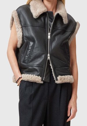 Kobieta w czarnej skórzanej kamizelce bez rękawów z beżowym futrzanym obszyciem i suwakiem, w połączeniu z ciemnymi spodniami w prążki i bransoletkami.