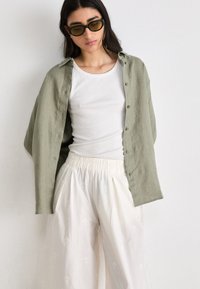 Chemise en lin vert olive à manches boutonnées sur un débardeur blanc, assortie à des pantalons plissés légers de couleur crème avec des motifs subtils.