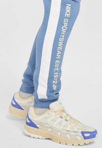 Světle modré tepláky s bílými bočními pruhy a žlutým nápisem "NIKE SPORTSWEAR EST.1972". Béžovo-modré tenisky s detaily z síťoviny.