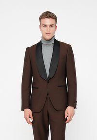 Tânăr îmbrăcat cu un costum maron cu rever din satin negru peste un pulover gri, stând în fața unui fundal alb simplu.