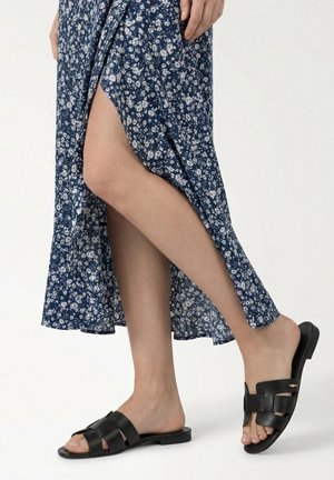 Nogi w niebieskiej sukience z kwiatowym wzorem i rozcięciem oraz czarnych płaskich sandałach typu slide z wyciętymi paskami na białym tle.