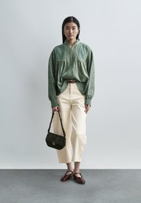 Γυναίκα που στέκεται φορώντας μια άνετη πράσινη μπλούζα, κρεμ παντελόνι, μπορντό παπούτσια Mary Jane, κρατώντας μια μαύρη τσάντα σε απλό γκρι φόντο.