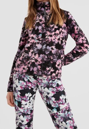 Kvinde iført en fleecepullover med lynlås i pink, sort og lilla blomsterprint samt matchende bukser med blomsterprint.
