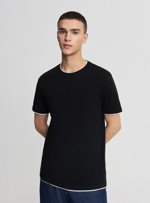 Młody mężczyzna z krótkimi włosami w czarnej koszulce z białym wykończeniem na rękawach, dekolcie i dolnym brzegu, stojący na tle jednolitego jasnego tła.