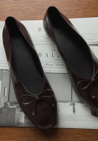 Hnedé kožené balerínky s hladkým povrchom, zaoblenou špičkou a mašľou na prednej strane, vystavené na tlačenom hotelovom písacom papieri.