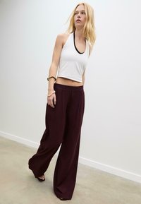 Žena kráčajúca vo vnútri oblečená v bielom bezrukávovom crop top, širokých tmavofialových nohaviciach a sandáloch na otvorenú špičku s náramkami.