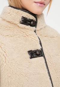 Veste en fausse fourrure beige avec un accent en cuir noir, dotée de deux boutons ronds et d'un col haut, mettant en valeur une texture douce et des lignes épurées.