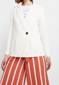 Bílý blejzr s jedním knoflíkem v želvovinovém stylu, se zářezovými klopami a dvěma předními kapsami. Nošený přes oranžovo-bílé pruhované široké kalhoty.