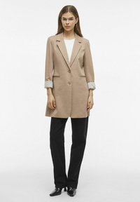 Blazer beige con un design sartorial, chiusura con un bottone singolo e dettagli a strisce sui polsini. Abbinato a pantaloni neri a gamba larga e scarpe a punta.