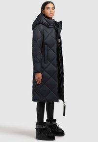Lange, donkerblauwe pufferjas met een ruitpatroon, hoge kraag, ritssluiting, zijzakken, gedragen over zwarte leggings en laarzen.