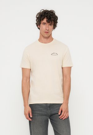 Young man with curly hair wearing a beige short-sleeve t-shirt with small black text on the left chest and gray jeans.