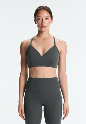 Kvinde, som står og vender fremad, iført en mørkegrå sportstop med tynde flettede stropper og matchende højtaljede leggings mod en ensfarvet baggrund.