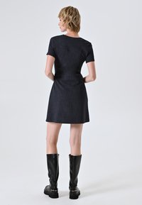 Kurzärmeliges Kleid aus dunklem Denim mit taillierter Passform und ausgestelltem Saum. Das Model kombiniert es mit kniehohen schwarzen Lederstiefeln. Minimalistisches Design.