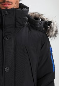 Homme portant une veste d'hiver noire avec de gros boutons, une capuche bordée de fourrure et une poche à fermeture éclair bleue sur la manche.