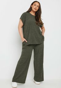 Oliivinvihreä kaksiosainen asu, jossa on löysä paita ja leveät housut. Rakeinen kangas ja taskut housuissa. Valkoiset tennarit jalassa.