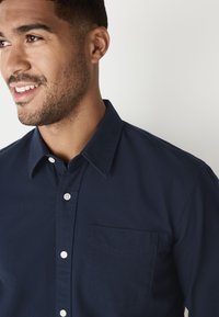 Marinblå bomullsskjorta med knappar fram, med spetsig krage, vita knappar och en bröstficka, har en slät textur.