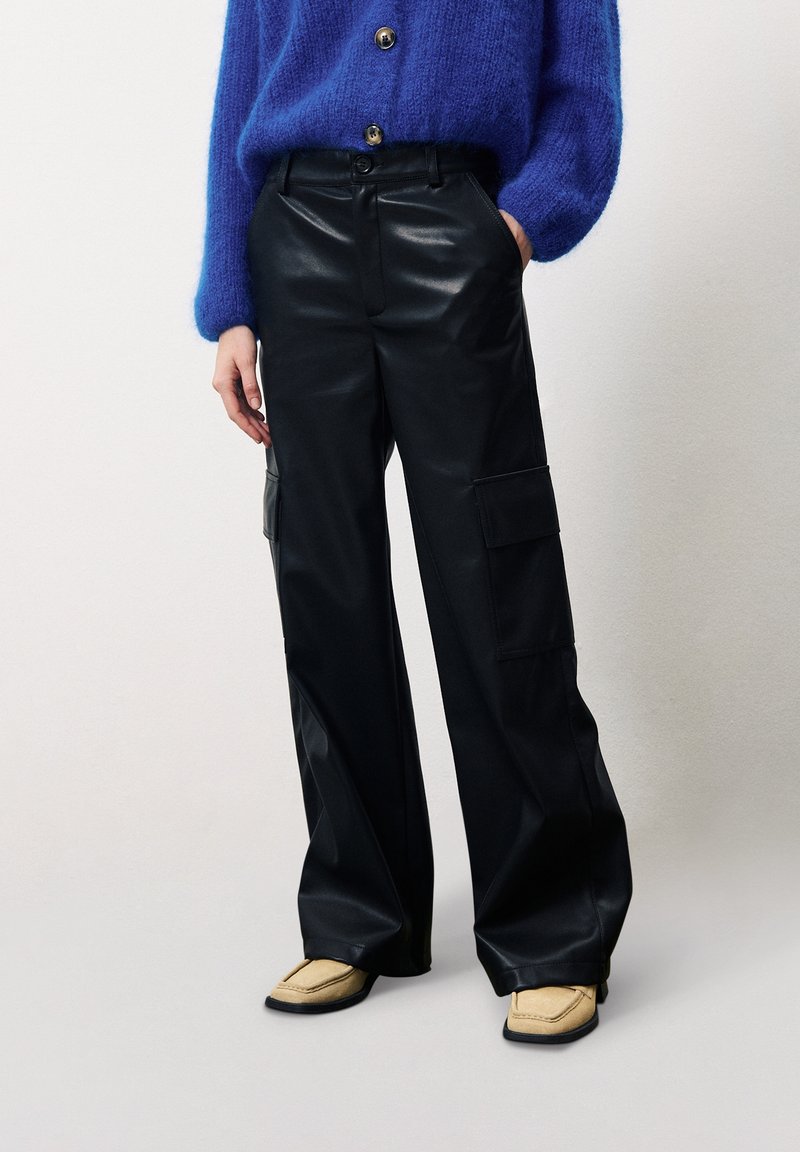 Pantalons cargo en simili cuir noir, à jambes larges, avec des poches latérales et une texture lisse. Associés à un pull bleu et des chaussures beiges.