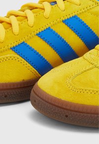 Zapatillas de ante amarillas con rayas de cuero azul, puntera redonda, suela de goma texturizada y costuras en la parte superior.