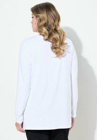 Langärmliges weißes Top mit relaxed Fit, glatter Textur und seitlichen Schlitzen, kombiniert mit schwarzen Hosen. Die Rückansicht zeigt einen geschwungenen Saum.