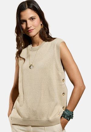Femme aux longs cheveux foncés portant un haut en tricot beige sans manches avec des boutons sur le côté, un collier avec pendentif en coquillage, et un bandana vert au poignet.