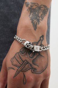 Zilveren kettingarmband met een schedelbedel en een sluiting, weergegeven op een hand met intrigerende tatoeages, tegen een donkere achtergrond.