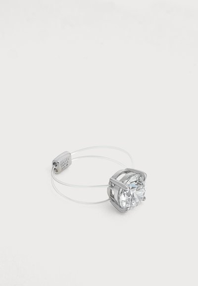 Anillo de plata con una gran piedra preciosa redonda y clara sujeta por cuatro garras y una banda delgada y transparente sobre un fondo blanco liso.