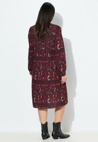 Langes, dunkles burgunderfarbenes Kleid mit floralem Muster, langen Ärmeln und smockierten Details. Getragen mit schwarzen Ankle Boots mit einem leichten Absatz.