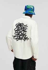 Camicia bianca a maniche lunghe con testo in stile graffiti nero sul retro. Il design presenta una texture liscia e polsini con bottoni. Abbinata a un cappello a secchiello con un motivo colorato.