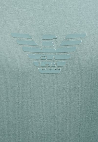Λογότυπο αετού με ανοιχτά φτερά πάνω από τα γράμματα "GA" σε φόν υφάσματος με υφή σε ανοιχτό μπλε χρώμα.