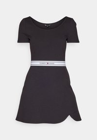 Mustat lyhythihaiset mekko puuvillasta, jossa on istuva vyötärö ja valkoinen nauha, jossa lukee "TOMMY JEANS", sekä kaareva helma yhdellä puolella.