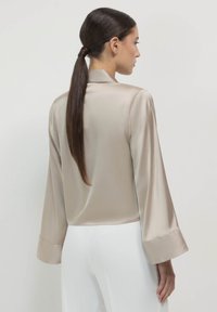 Blusa di raso beige con maniche lunghe e polsini larghi. Presenta una forma dritta e una texture morbida, mostrata di spalle con un'acconciatura a coda di cavallo.