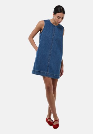 Femme debout, la tête inclinée vers le bas, portant une robe en denim bleu sans manches et des chaussures Mary Jane rouges, sur un fond clair uni.