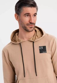 Beige hoodie med en svart logotyp-patch, dragsko med vita accenter och en framficka. Mjuk tyg och avslappnad passform.