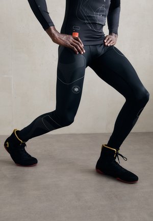Schwarzes Sportoutfit mit strukturierten Einsätzen und weißen Akzenten, kombiniert mit schwarzen High-Top-Schuhen mit gelben Details und roten Sohlen.