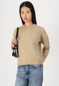 Maglione beige lavorato a maglia con un motivo testurizzato, scollatura rotonda e polsini a costine. Abbinato a jeans attillati azzurri e una piccola borsa nera.