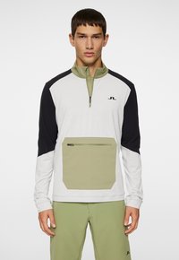 Quarter-zip trøje i hvid, sort og olivengrøn. Har en stor forlommes og et logo på brystet. Glat stofstruktur.