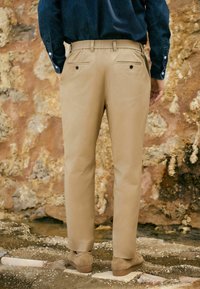 Beige Chinos mit einem entspannten Schnitt, elastischem Bund und Gesäßtaschen. Glattes Material mit minimaler Textur, kombiniert mit braunen Wildlederschuhen.