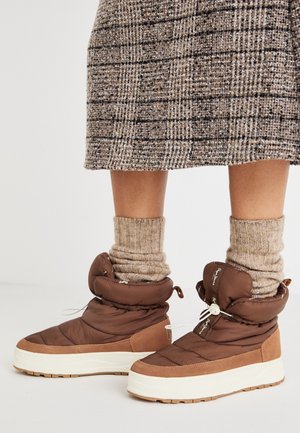 Botas de tobillo aisladas marrones con un upper suave y acolchado y un cordón decorativo, asentadas sobre una suela de plataforma de goma blanca. Calcetines beige texturizados visibles.