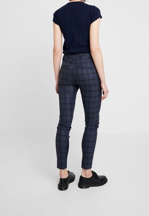 Kobieta w granatowym topie z krótkim rękawem, obcisłych ciemnoniebieskich spodniach w kratę oraz czarnych sznurowanych butach, stojąca na tle jednolitego jasnego tła.