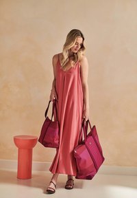 Long pink dress with spaghetti straps, paired with two pink striped tote bags. Background features a smooth, light-toned wall and a round stool.