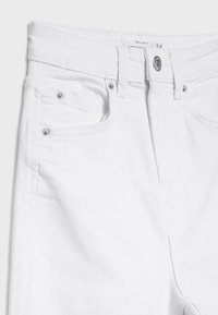 Jean en denim blanc avec fermeture par boutons, passants pour ceinture, poches avant et poche monnaie avec rivets métalliques, gros plan sur la partie avant supérieure.