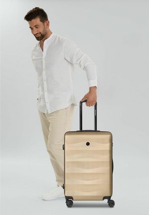 Homme en chemise blanche et pantalon beige tirant une valise rigide dorée avec poignée noire et quatre roues sur un fond gris.