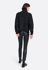 Schwarzer Strickpullover mit Rollkragen in lockerer Passform, kombiniert mit enganliegenden schwarzen Lederhosen und schwarzen Schuhen, von hinten betrachtet.