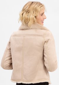 Lächelnde Person mit kurzen lockigen blonden Haaren, die eine beige Schafwolljacke trägt, von hinten betrachtet und leicht nach rechts gewandt.