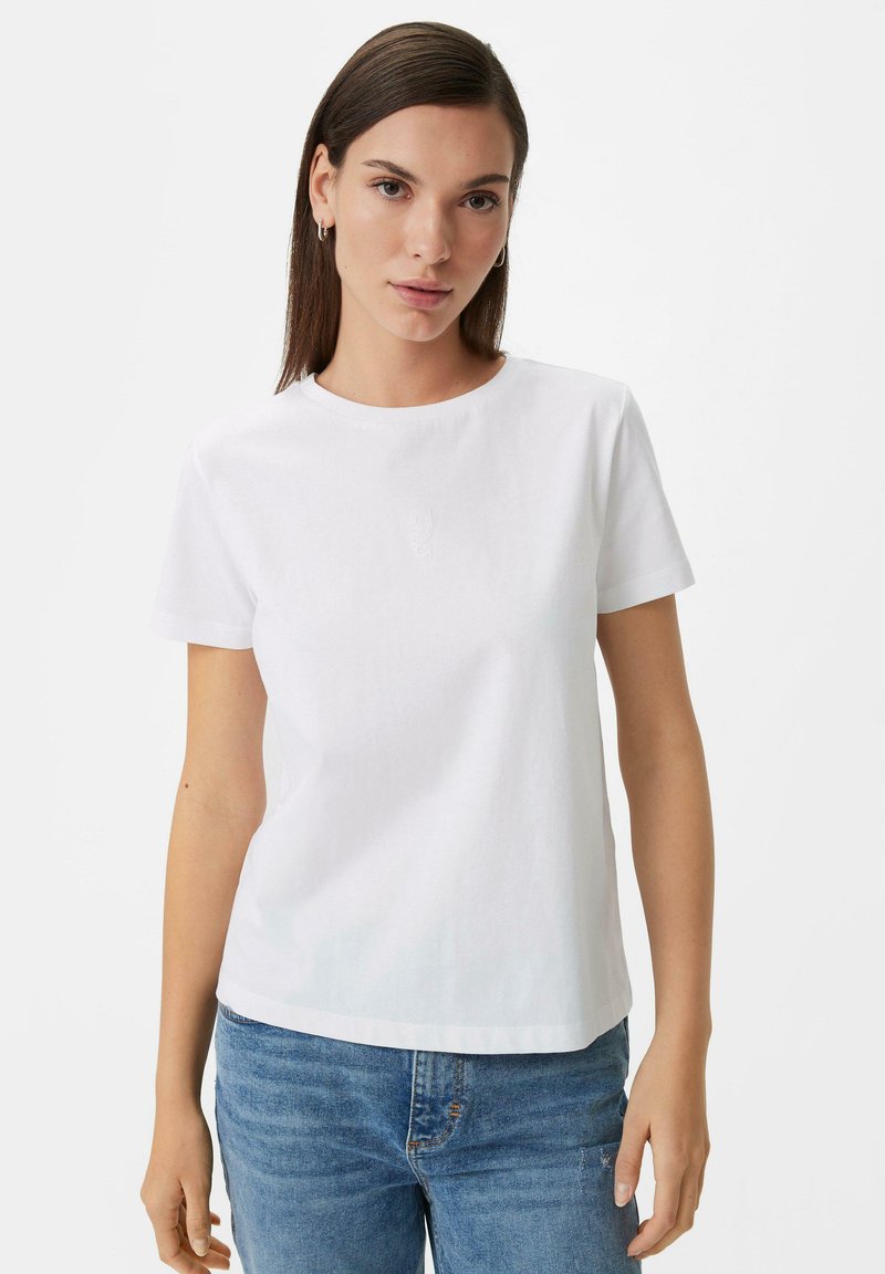 White cotton t-shirt, short sleeves, round neckline, subtly embossed logo on the chest, paired with blue denim jeans.